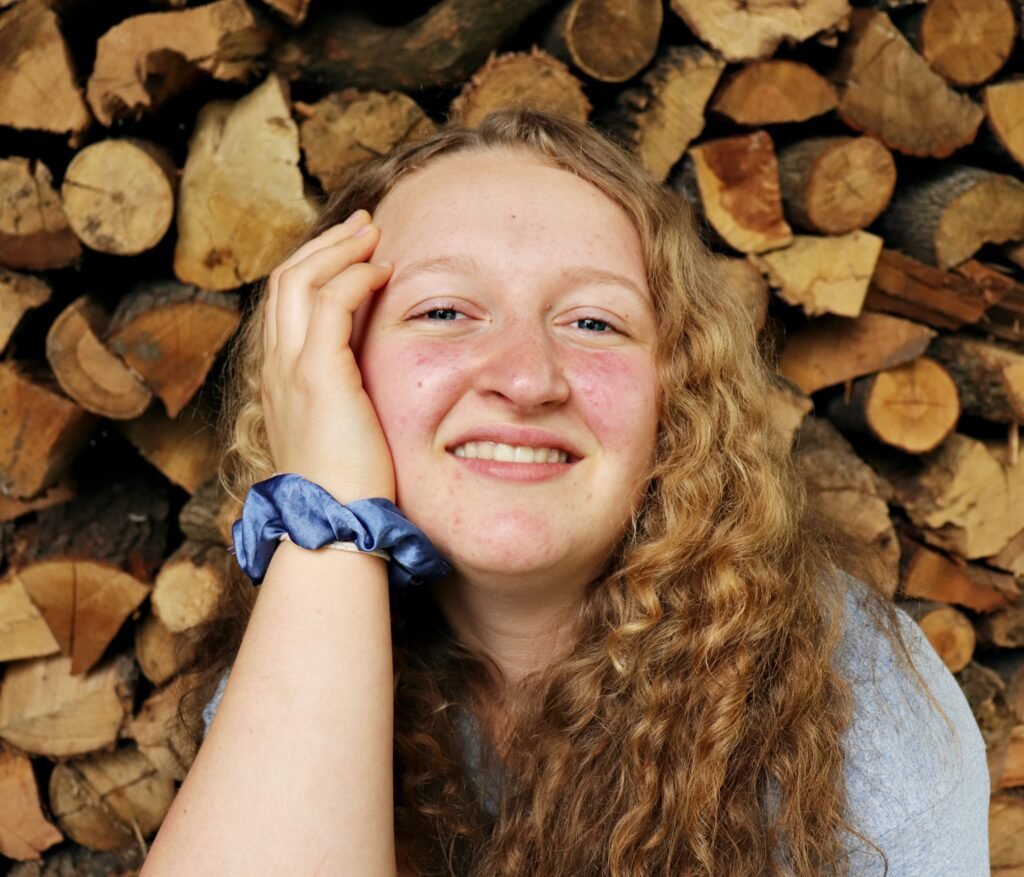 Photo of surface pattern designer 
and artist Noemi Salome. She has blue eyes, blond-brown, curly hair, and a large smile. She is posed in front of a wall of stacked firewood.